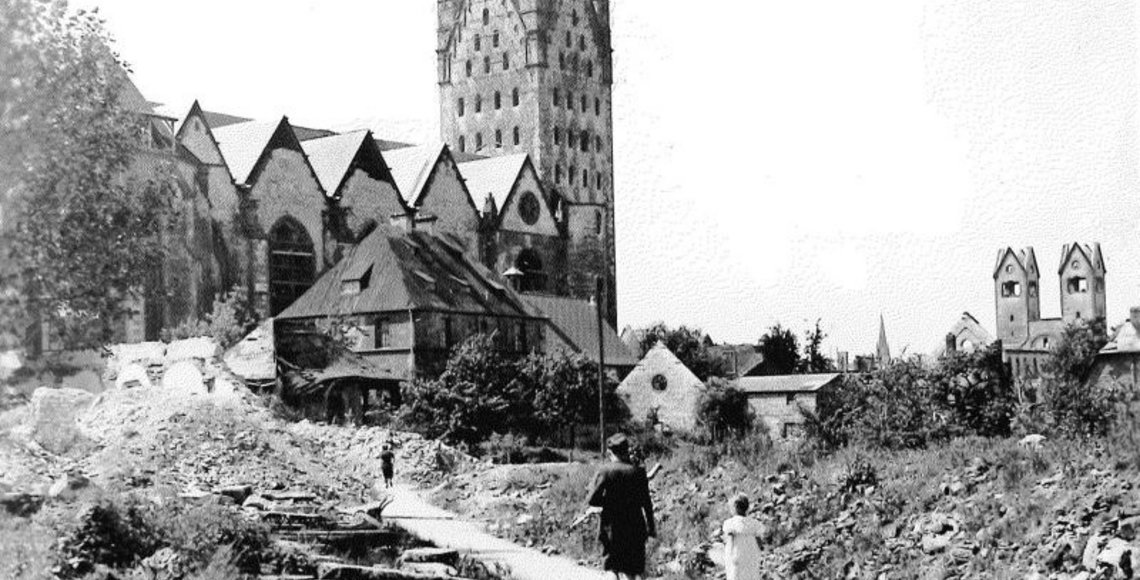 Das Foto von 1949 zeigt im Vordergrund eine Mutter mit Kind, die durch das durch den Zweiten Weltkrieg massiv zerstörte Paderborn auf den Dom zuläuft, der ebenfalls zum Teil zerstört wurde.